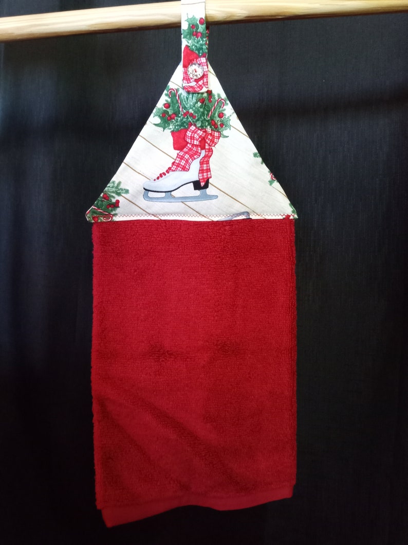 K&ouml;nnte beinhalten: Rotes K&uuml;chentuch mit einer wei&szlig;en und gr&uuml;nen Schleife aus Weihnachtsstoff. Die Schleife zeigt einen wei&szlig;en Schlittschuh mit einer roten Schleife und Stechpalme.