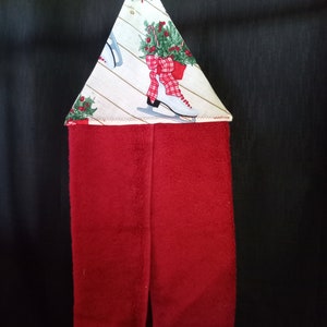 K&ouml;nnte beinhalten: Ein rotes K&uuml;chentuch mit einer wei&szlig;en und gr&uuml;nen, festlichen Stoffschlaufe zum Aufh&auml;ngen. Die Schlaufe hat einen wei&szlig;en Hintergrund mit einer rot-wei&szlig; karierten Schleife, gr&uuml;nem Stechpalmenlaub und wei&szlig;en Schlittschuhen.