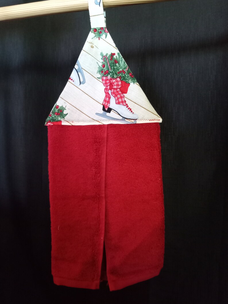 K&ouml;nnte beinhalten: Ein rotes K&uuml;chentuch mit einer wei&szlig;en und gr&uuml;nen, festlichen Stoffschlaufe zum Aufh&auml;ngen. Die Schlaufe hat einen wei&szlig;en Hintergrund mit einer rot-wei&szlig; karierten Schleife, gr&uuml;nem Stechpalmenlaub und wei&szlig;en Schlittschuhen.