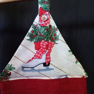 K&ouml;nnte beinhalten: Ein rot-wei&szlig;es Weihnachts-K&uuml;chenhandtuch mit einer wei&szlig;en Schlittschuh mit einer roten Schleife und einem Stechpalmen-Design. Das Handtuch hat eine Schlaufe zum Aufh&auml;ngen und einen roten Knopf.