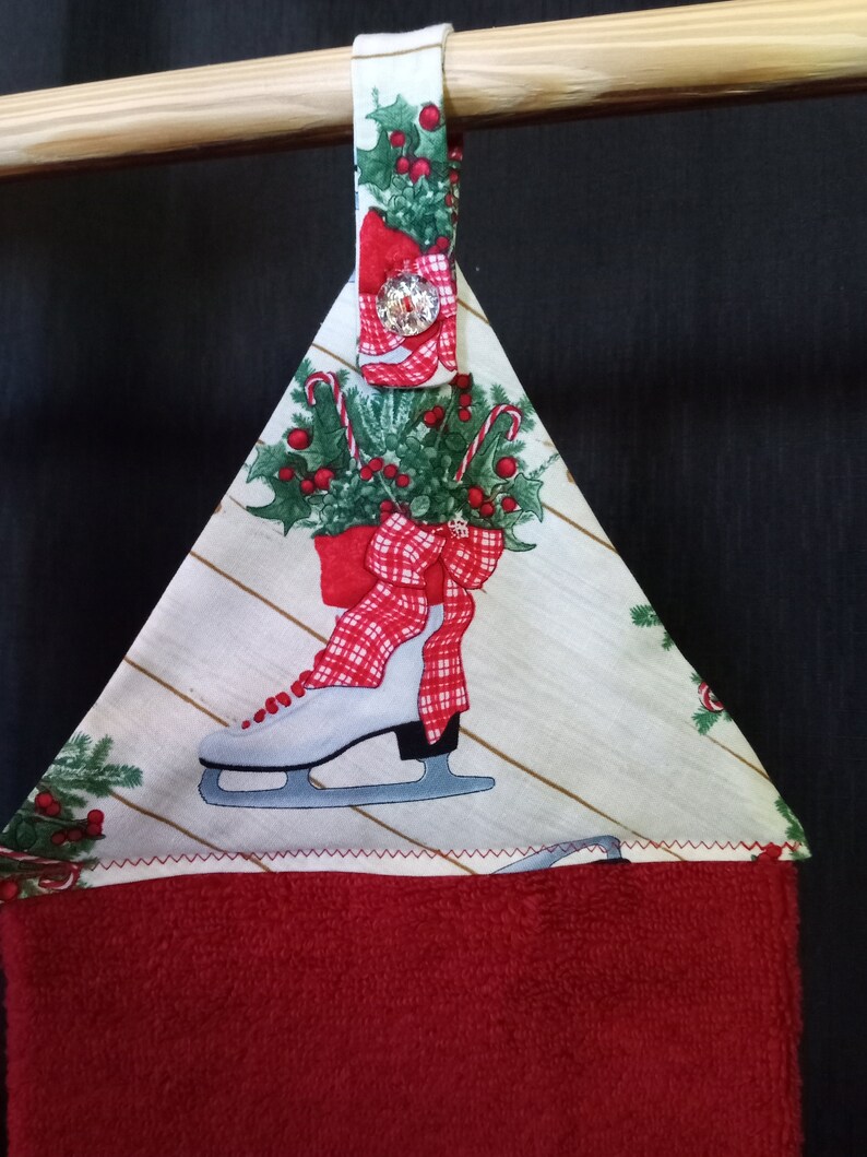 K&ouml;nnte beinhalten: Ein rot-wei&szlig;es Weihnachts-K&uuml;chenhandtuch mit einer wei&szlig;en Schlittschuh mit einer roten Schleife und einem Stechpalmen-Design. Das Handtuch hat eine Schlaufe zum Aufh&auml;ngen und einen roten Knopf.