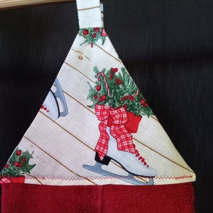 K&ouml;nnte beinhalten: Ein rot-wei&szlig;es Geschirrtuch mit Weihnachtsmotiv, wei&szlig;em Hintergrund und rotem Rand. Das Tuch zeigt ein Paar Schlittschuhe mit einer roten Schleife und einem gr&uuml;nen Stechpalmenzweig. Das Tuch hat eine Schlaufe zum Aufh&auml;ngen.