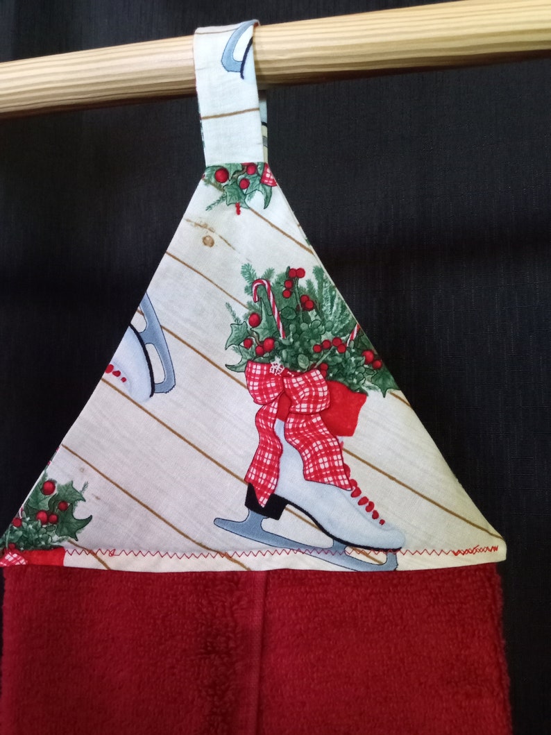 K&ouml;nnte beinhalten: Ein rot-wei&szlig;es Geschirrtuch mit Weihnachtsmotiv, wei&szlig;em Hintergrund und rotem Rand. Das Tuch zeigt ein Paar Schlittschuhe mit einer roten Schleife und einem gr&uuml;nen Stechpalmenzweig. Das Tuch hat eine Schlaufe zum Aufh&auml;ngen.