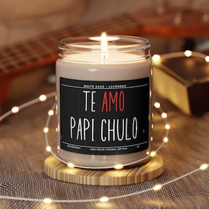 May include: A brown soy candle with a black label that reads "WHITE SAGE LAVENDER TE AMO PAPI CHULO" in white text with "AMO" in red text. The candle is on a wooden coaster and surrounded by white string lights.