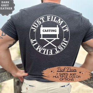 May include: A dark grey heather t-shirt with a white circle graphic on the back. The graphic features a director's chair with the word "CASTING" on the back and the text "JUST FILM IT" circling around the chair.