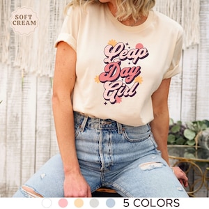 May include: A light beige t-shirt with a retro-style graphic that says "Leap Day Girl" in pink and orange lettering with flowers and stars. The shirt is being worn by a person with blonde hair and blue jeans.