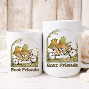 May include: Two green frogs riding a tandem bicycle in a forest setting. The mugs are white with the image printed on them. The text "Best Friends" is printed below the image.