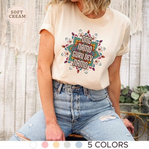 May include: A cream colored t-shirt with a mandala design featuring the text "Ong Namo Guru Dev Namo". The shirt is shown on a model wearing blue jeans.
