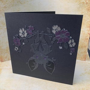 May include: A square black greeting card featuring a die-cut illustration of a bat hanging upside down from a branch. The bat has purple eyes and is surrounded by dark purple and grey floral accents with small purple gems embedded in the flowers.