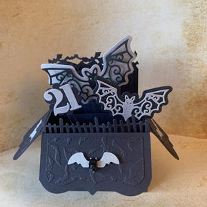 May include: A black paper pop-up box card featuring intricate silver bat cutouts, a fence border, and the number 21 in silver. The front panel is embossed with a bat pattern and decorated with a small silver bat and a black skull embellishment.