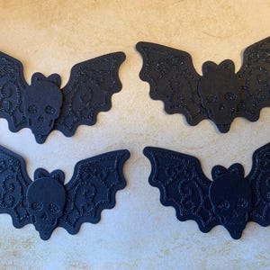 May include: Four black bat decorations with skull faces. The bats feature detailed wing patterns and are covered in glitter. Ideal for Halloween, these decorations add a spooky touch to crafts or parties. Each bat is approximately 15 cm wide.
