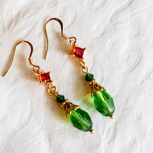 May include: A pair of gold-coloured dangle earrings. Each earring features a red square gem, green beads, and a teardrop-shaped green bead. The earrings are set against a white background.