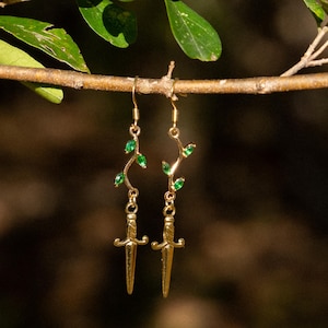 May include: Gold earrings with a delicate vine design and emerald green stones. The vine ends with a dagger shape.