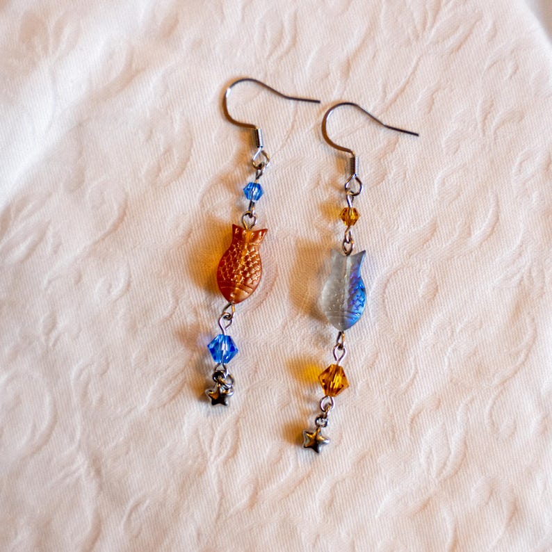 May include: A pair of dangle earrings featuring fish-shaped charms in orange and blue hues. The earrings have silver-tone hooks and are adorned with blue and amber-colored beads, and small star charms. The earrings are displayed on a cream-colored surface.