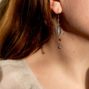 May include: A silver earring with a gray, teardrop-shaped bead, amber-colored crystals, and a small star charm. The earring is suspended from a delicate silver hook. The earring is shown against a blurred background.