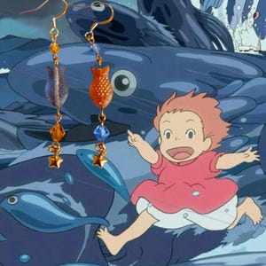 May include: Gold-colored dangle earrings featuring fish-shaped and star-shaped charms. The earrings have orange, blue, and clear beads. The background is a cartoon scene with a child running and blue fish.