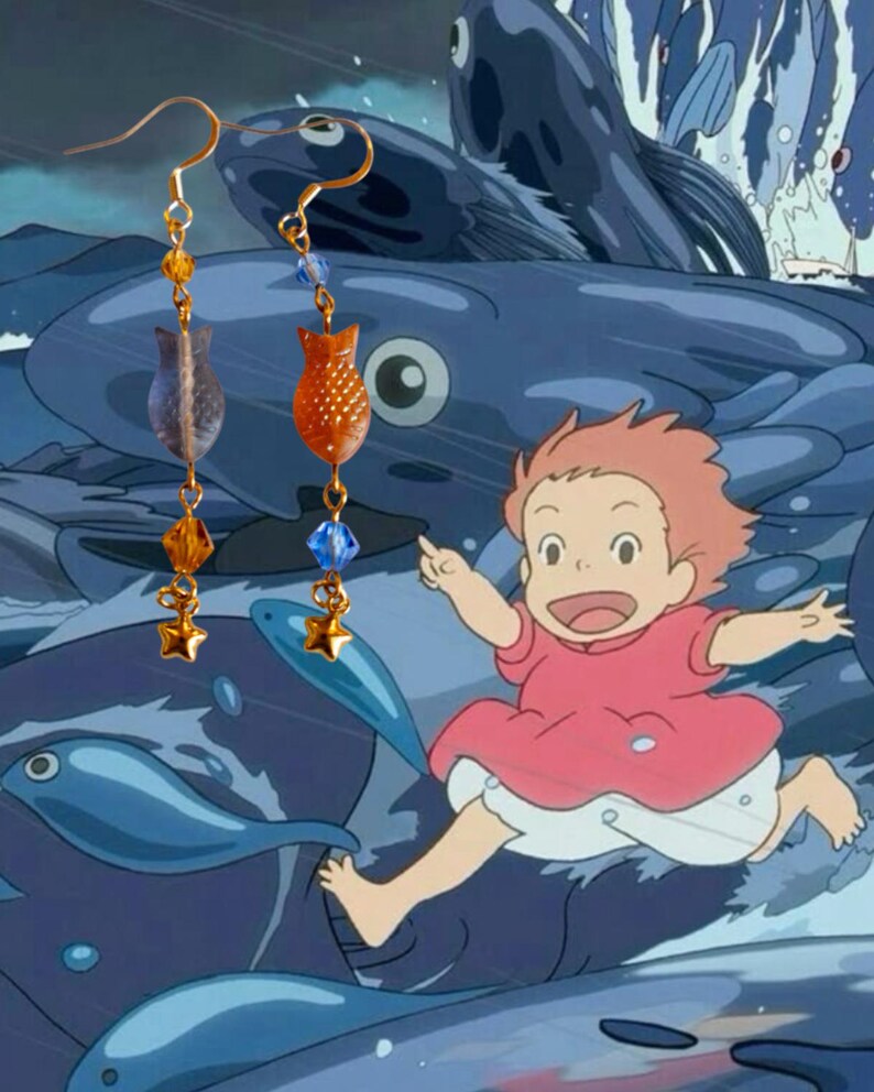 May include: Gold-colored dangle earrings featuring fish-shaped and star-shaped charms. The earrings have orange, blue, and clear beads. The background is a cartoon scene with a child running and blue fish.