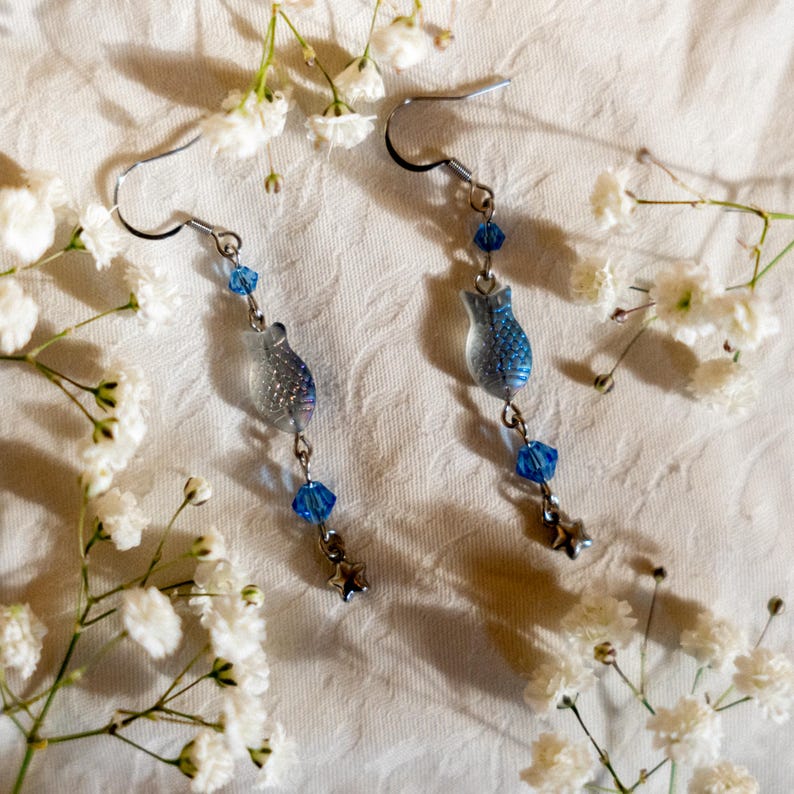 May include: A pair of dangle earrings with a fish-shaped design. The earrings feature a silver-tone metal hook, blue beads, and a clear, textured fish pendant with blue glitter accents. A small star charm hangs at the bottom of each earring.