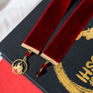 Puede incluir: Dos marcapáginas de terciopelo rojo con ribetes dorados y un dije dorado con un diseño de llama. Los dijes están unidos a los marcapáginas con cuentas de cristal rojo.