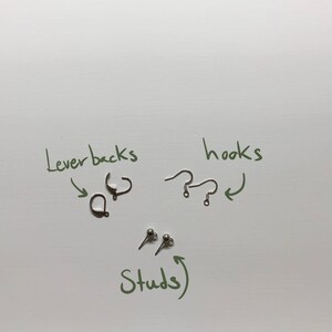May include: An assortment of earring findings, including lever backs, hooks, and stud earrings, are displayed on a white surface. The image includes handwritten labels pointing to each type of earring component. The components are in silver and bronze tones.