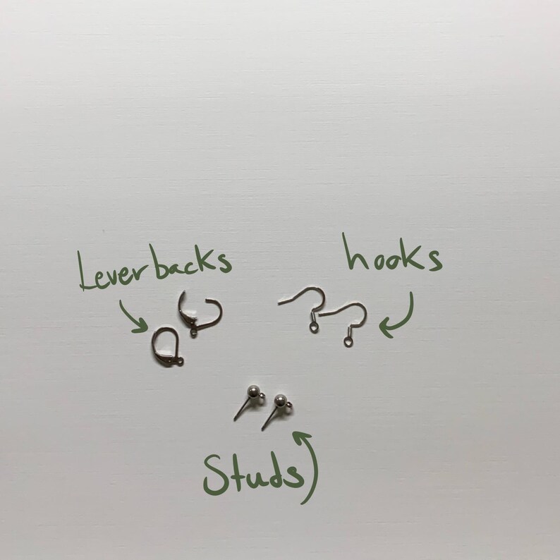 May include: An assortment of earring findings, including lever backs, hooks, and stud earrings, are displayed on a white surface. The image includes handwritten labels pointing to each type of earring component. The components are in silver and bronze tones.