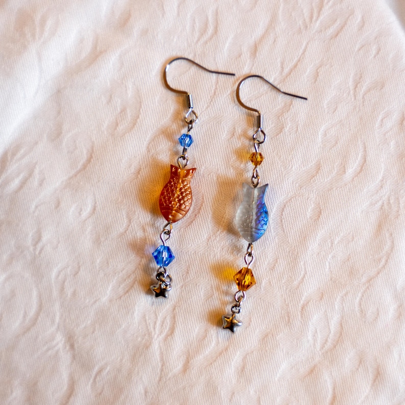 May include: A pair of dangle earrings featuring fish-shaped charms in orange and clear blue. The earrings have blue and yellow beads, and small star charms. The hooks are silver-colored.