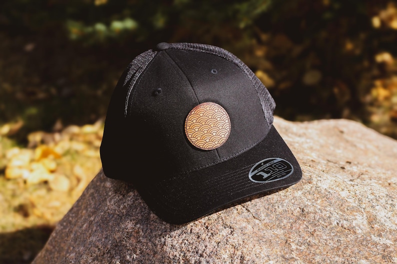 May include: Black mesh baseball cap with a black brim and a round, brown leather patch with a wave pattern.