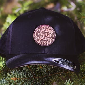 May include: A black mesh trucker hat with a brown leather patch featuring a red and white wave pattern.