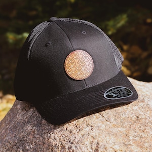 May include: A black baseball cap with a brown leather patch featuring a geometric pattern. The cap has a mesh back and a curved brim.