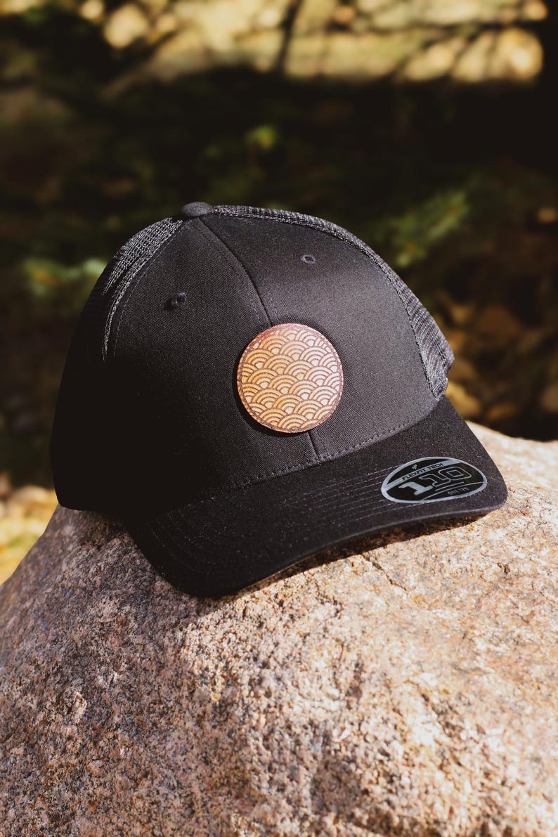 May include: A black baseball cap with a brown leather patch featuring a geometric pattern. The cap has a mesh back and a curved brim.