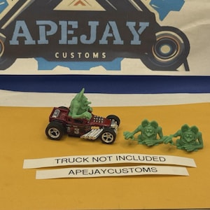 May include: A maroon die-cast toy car with a green monster figure seated inside, accompanied by two additional green monster figures. The text APEJAY CUSTOMS is visible at the top, and TRUCK NOT INCLUDED and APEJAYCUSTOMS are printed on white strips.