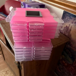 May include: A pink plastic organizer with multiple clear, lidded compartments stacked in a cube shape. The organizer sits on a wooden surface, with a dark gray square in the center of the top.