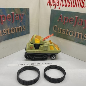 May include: A vintage olive green toy tank with orange translucent windows and a top-mounted orange cannon. The vehicle features black treads and the number 2000 printed on the side. Two separate black rubber treads are placed in the foreground.