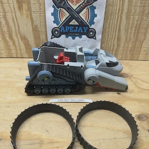 May include: A gray plastic toy tank vehicle with black treads and a red interior component, accompanied by two separate black circular rubber treads. A white paper sign displays the text APEJAY CUSTOMS with a wrench and brush logo.