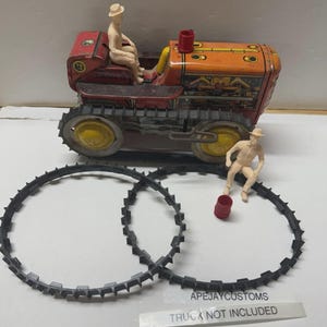 May include: A vintage-style red and orange metal toy tractor with black rubber tracks, featuring two cream-coloured plastic figures wearing hats, a small red plastic barrel, and a label reading APEJAYCUSTOMS TRUCK NOT INCLUDED.