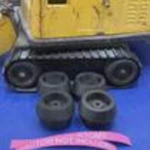 May include: A yellow toy excavator with black tracks positioned behind four small, dark gray circular plastic wheels. Two curved pink strips of paper with white text are placed in the foreground, partially obscuring the view of the base.