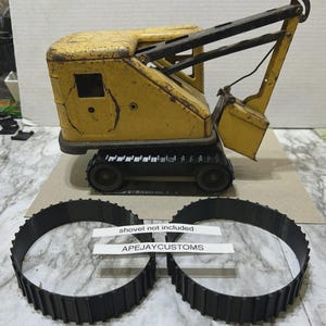 May include: A yellow and black metal toy crane with a bucket and crawler tracks. Two separate black replacement tracks are positioned in front. Two white paper labels are present with the text: shovel not included and APEJAYCUSTOMS.