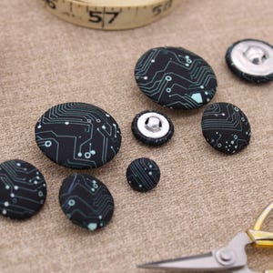 May include: Assortment of black and green fabric buttons with a circuit board design. The buttons are various sizes and are arranged on a brown fabric surface.