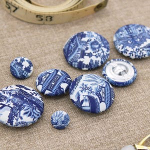 May include: A selection of blue and white fabric-covered buttons in different sizes. The buttons display a detailed, patterned design. A measuring tape is visible in the background, suggesting a sewing or craft theme.