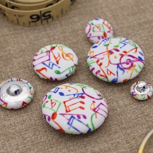 May include: A selection of fabric-covered buttons in various sizes, patterned with colourful musical notes on a white background. The buttons are scattered on a textured surface, with a measuring tape visible in the background.