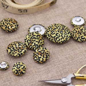 May include: A collection of eight yellow and black leopard print fabric buttons. The buttons are various sizes and are arranged on a beige fabric surface.