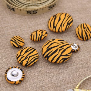 May include: A set of seven tiger stripe patterned buttons in various sizes. The buttons are made of fabric and have a black and orange color scheme.