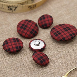 May include: A selection of red and black tartan fabric-covered buttons in different sizes. One button has a silver metal back. The buttons are on a beige surface, with a measuring tape and a gold object in the background.