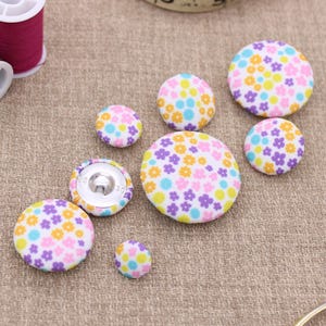 May include: A selection of fabric-covered buttons in different sizes, decorated with a floral pattern in pastel shades. The buttons have a metal shank for sewing. Spools of thread are also visible.