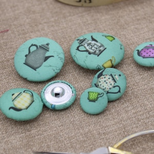 Può includere: Una collezione di bottoni rivestiti in tessuto color teal con vari disegni di teiere. I bottoni variano nelle dimensioni e presentano diverse illustrazioni di teiere in nero, bianco e rosa. Un bottone ha un retro a scatto argentato. Forbici e filo sullo sfondo.