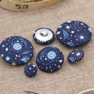 May include: A selection of fabric-covered buttons in different sizes. The buttons display a dark blue celestial design with white stars and swirling patterns. One button has a silver metal back. The buttons are arranged on a neutral-toned surface.