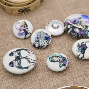 May include: A collection of fabric-covered buttons with illustrations from Alice in Wonderland. The buttons feature characters like Alice, the White Rabbit, and the Mad Hatter, in various sizes and designs. A measuring tape is in the background.