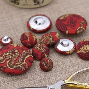 May include: A collection of red fabric-covered buttons with a gold dragon and pattern design. The buttons vary in size and have silver metal backs. A pair of gold-handled scissors is in the foreground.