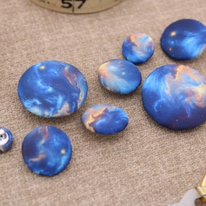 May include: A collection of fabric-covered buttons in various sizes, featuring a vibrant blue and gold galaxy print. The buttons are arranged on a neutral-toned fabric surface, with a spool of red thread and a pair of scissors visible in the background.