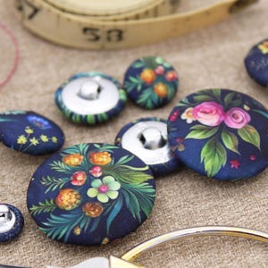 May include: Navy blue fabric buttons with floral patterns in pink, orange, and green. Some buttons have a silver metal shank. A measuring tape and scissors are in the background, suggesting a sewing or craft theme.